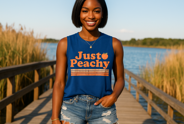 AI Model : A woman with deep brown skin and short, straight black hair stands on a wooden boardwalk at a lakeside dock, wearing a royal blue “Just Peachy” sleeveless tank and distressed denim shorts. Wild reeds and sparkling water surround her under a bright, clear sky.