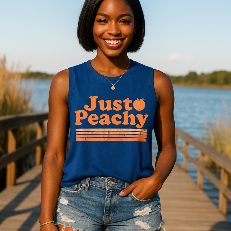 AI Model : A woman with deep brown skin and short, straight black hair stands on a wooden boardwalk at a lakeside dock, wearing a royal blue “Just Peachy” sleeveless tank and distressed denim shorts. Wild reeds and sparkling water surround her under a bright, clear sky.